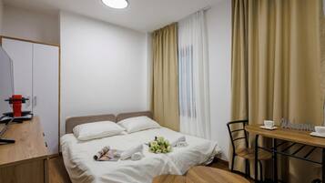 A hotel room with a bed, a desk with a computer, a chair, a table with a 'Welcome' sign, and a window with curtains.