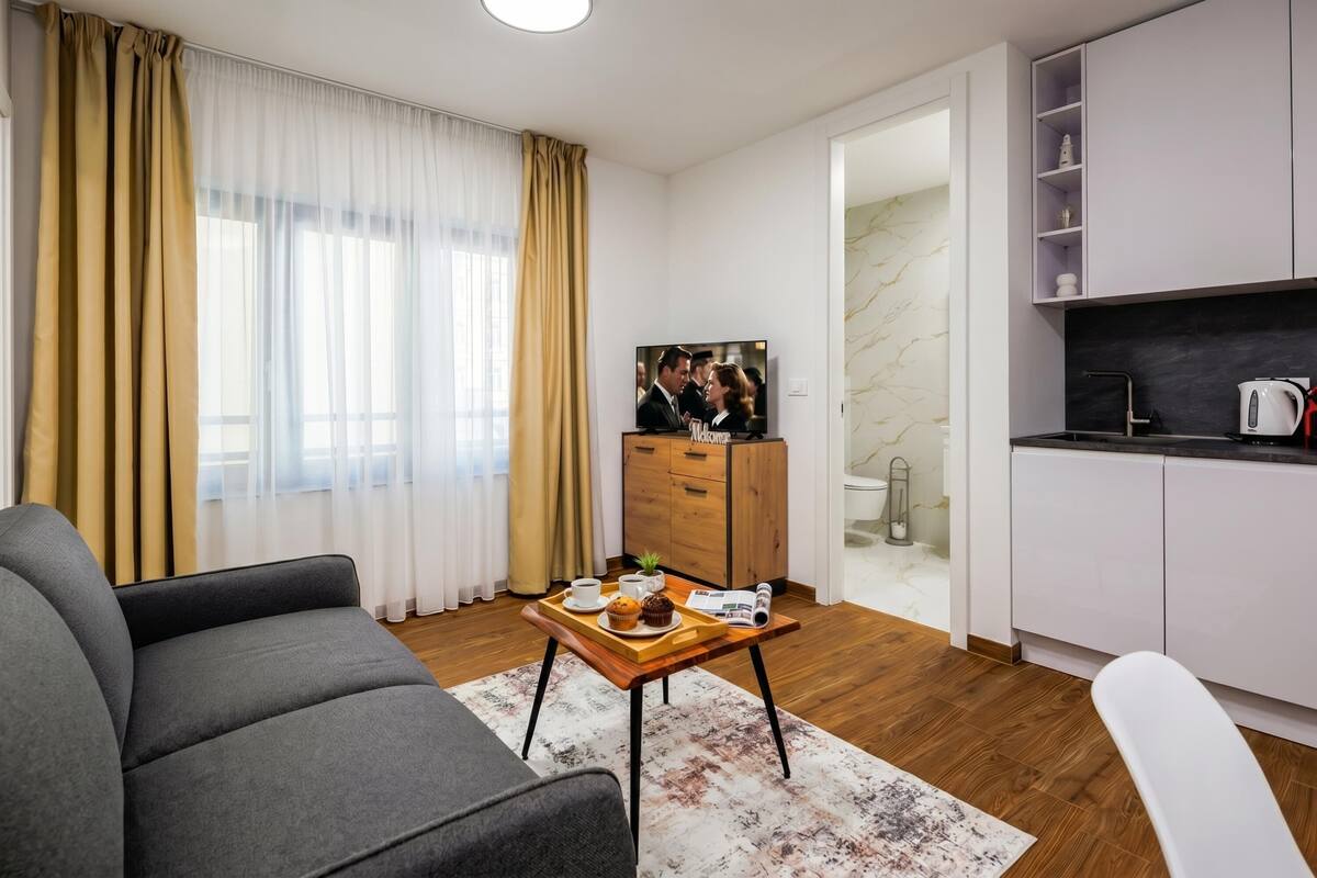 A modern living room with a sofa, a coffee table with a tray of food, a TV, and a kitchen area with cabinets and a kettle.