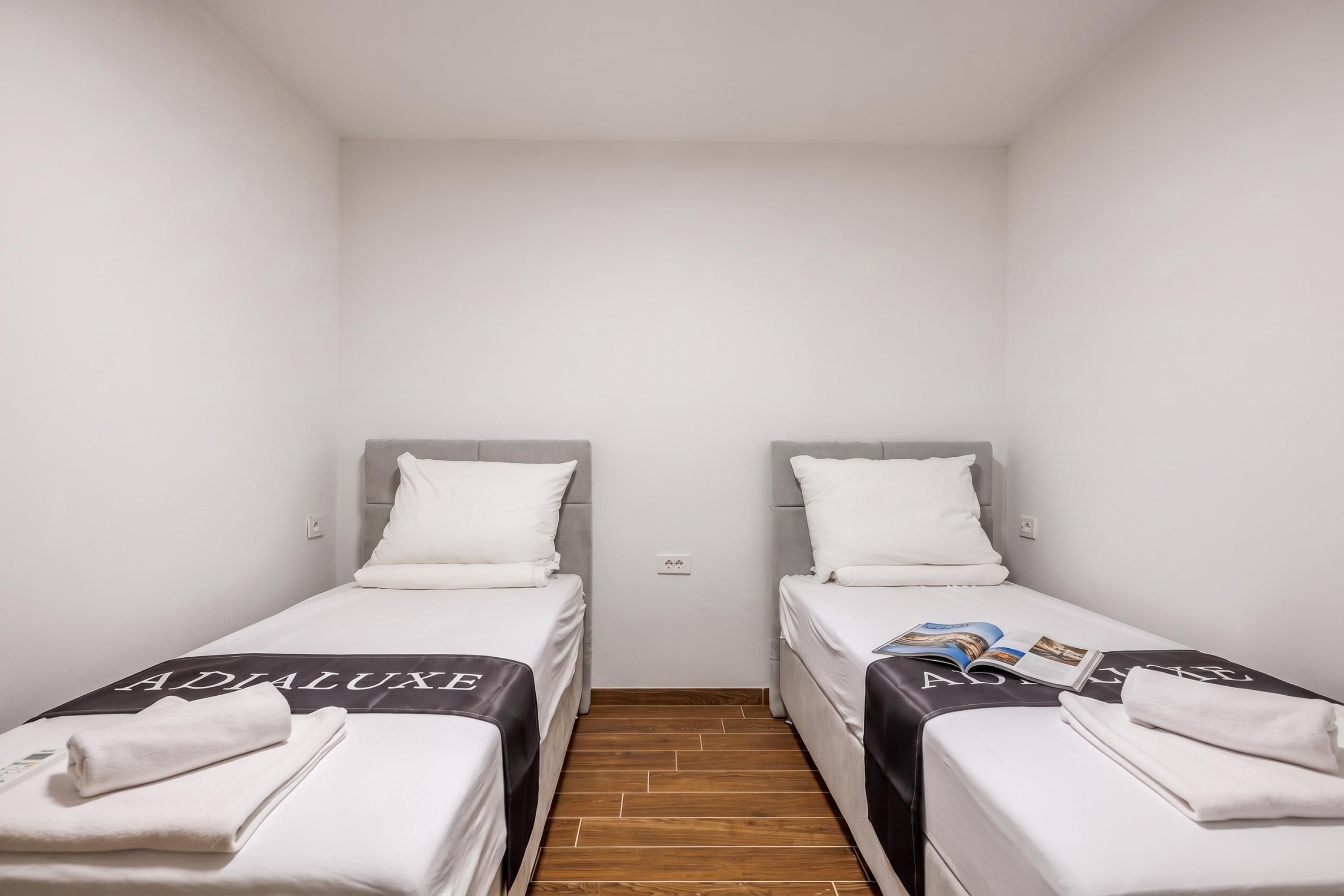 Une petite chambre propre, avec deux lits simples, chacun doté d’une tête de lit grise et de draps blancs.