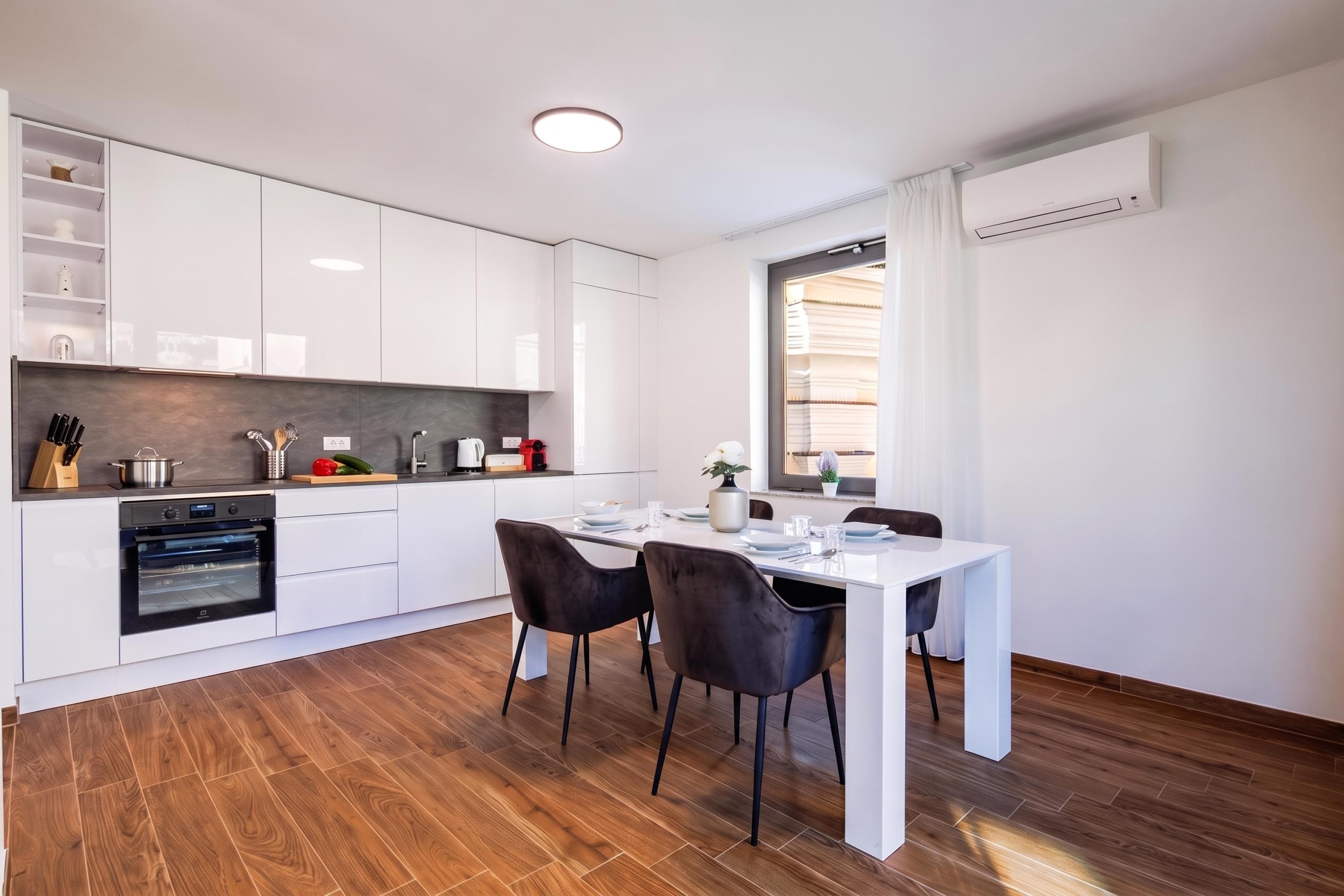 A modern kitchen with white cabinetry, a built-in oven, and a dining area with a white table and two chairs.