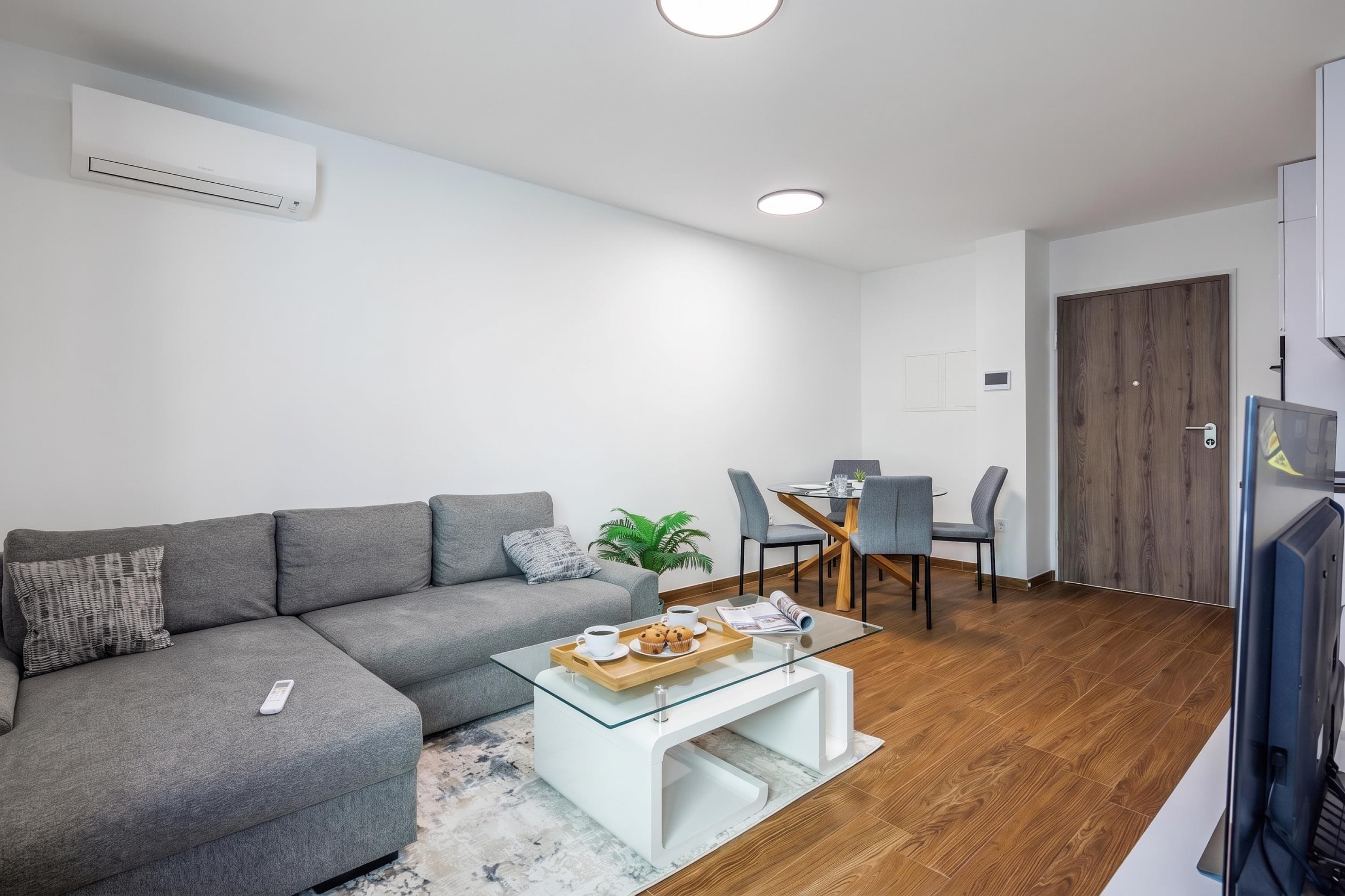 A modern living room with a grey sofa, a glass coffee table, and a dining area with a wooden table and chairs.