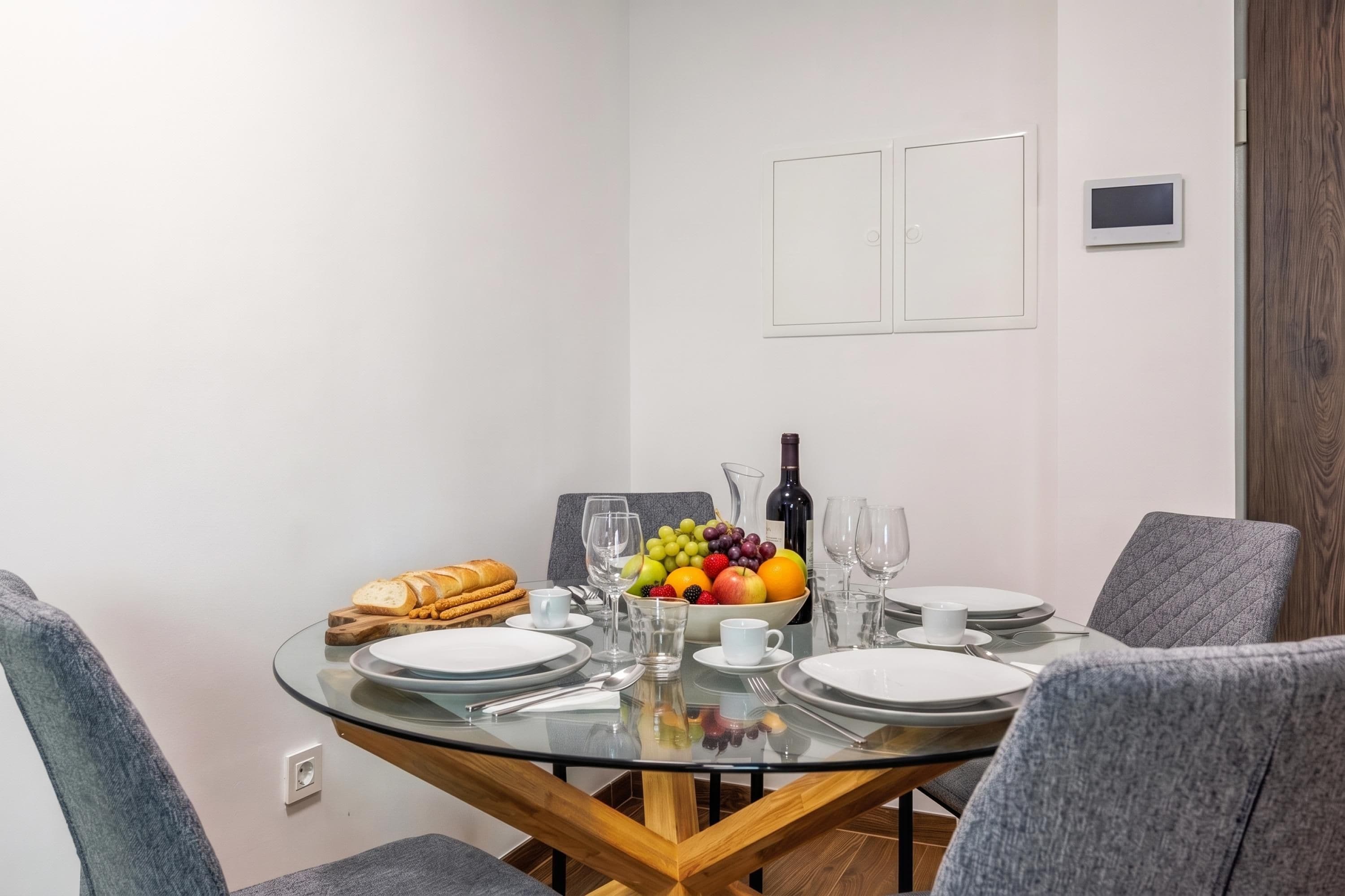A dining area with a round glass table, grey upholstered chairs, and a fruit bowl with various fruits and a bottle of wine.
