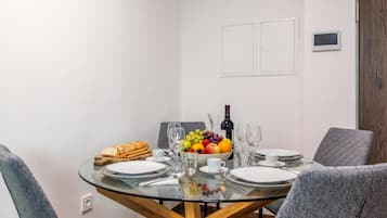 A dining area with a round glass table, grey upholstered chairs, and a fruit bowl with various fruits and a bottle of wine.