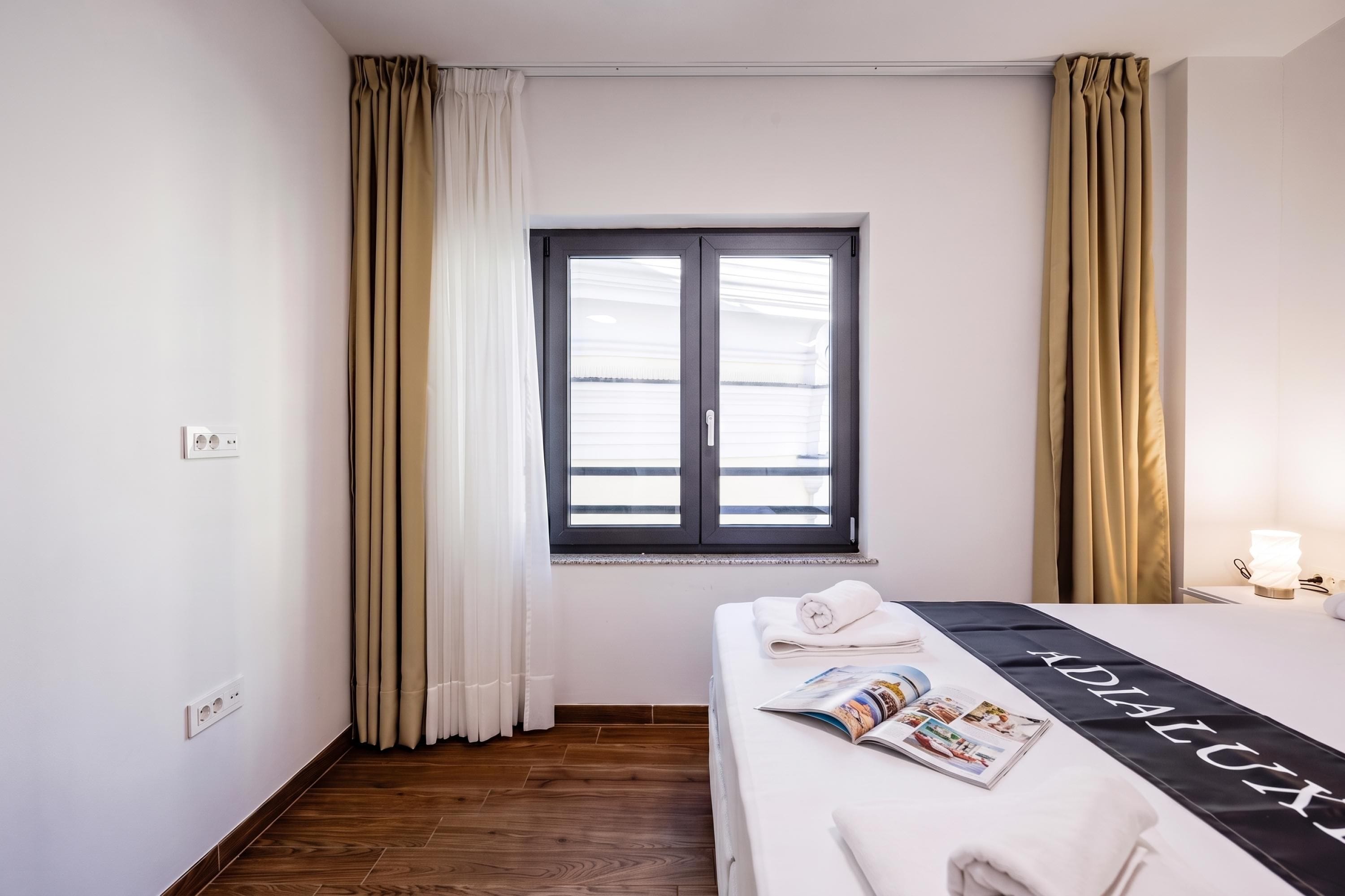 Ett hotellrum med en säng, ett fönster med gardiner, ett nattduksbord med en lampa och en tidning på sängen.