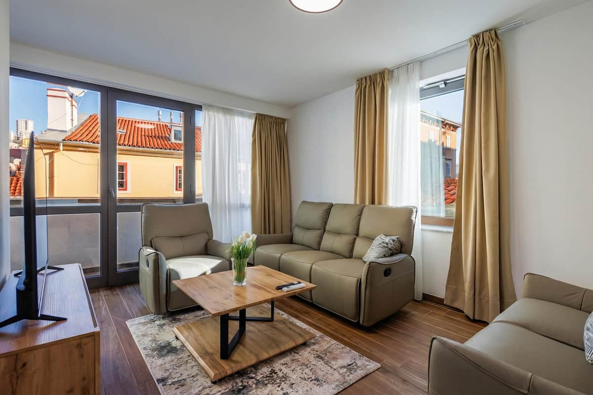 A modern living room with a sofa, armchairs, a coffee table, and a TV. Large windows offer a view of buildings outside.