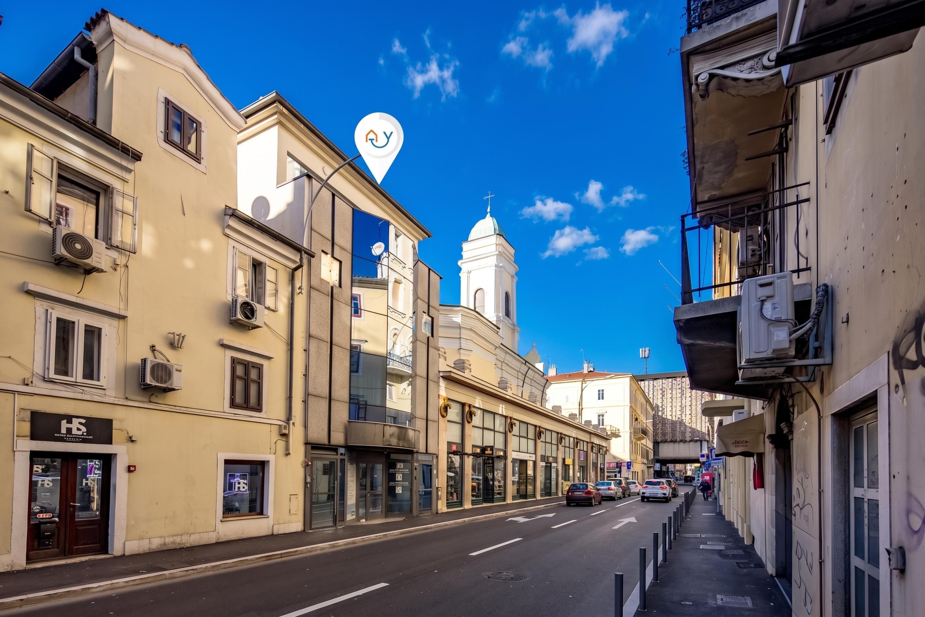 Vista da rua com edifícios, céu claro e uma torre de igreja imponente.