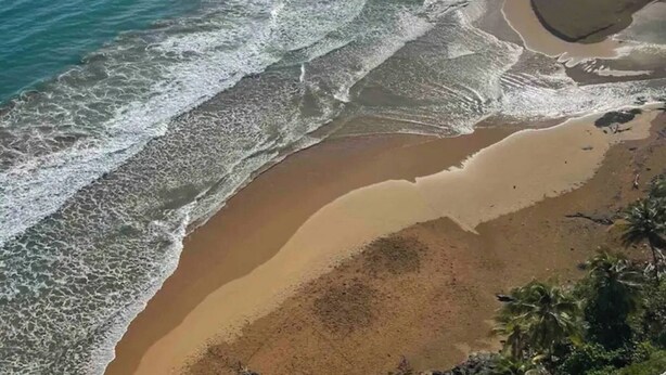A coastal beach scene with waves reaching the shore, a clear sky, and distant land. - DUERMES CON LA NATURALEZA (Ramos)