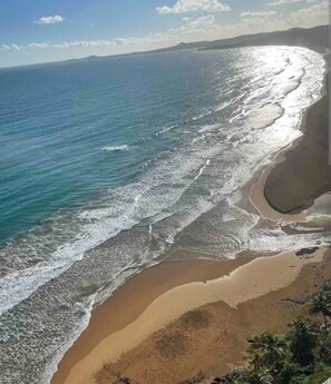 A coastal beach scene with waves reaching the shore, a clear sky, and distant land. - DUERMES CON LA NATURALEZA (Ramos)