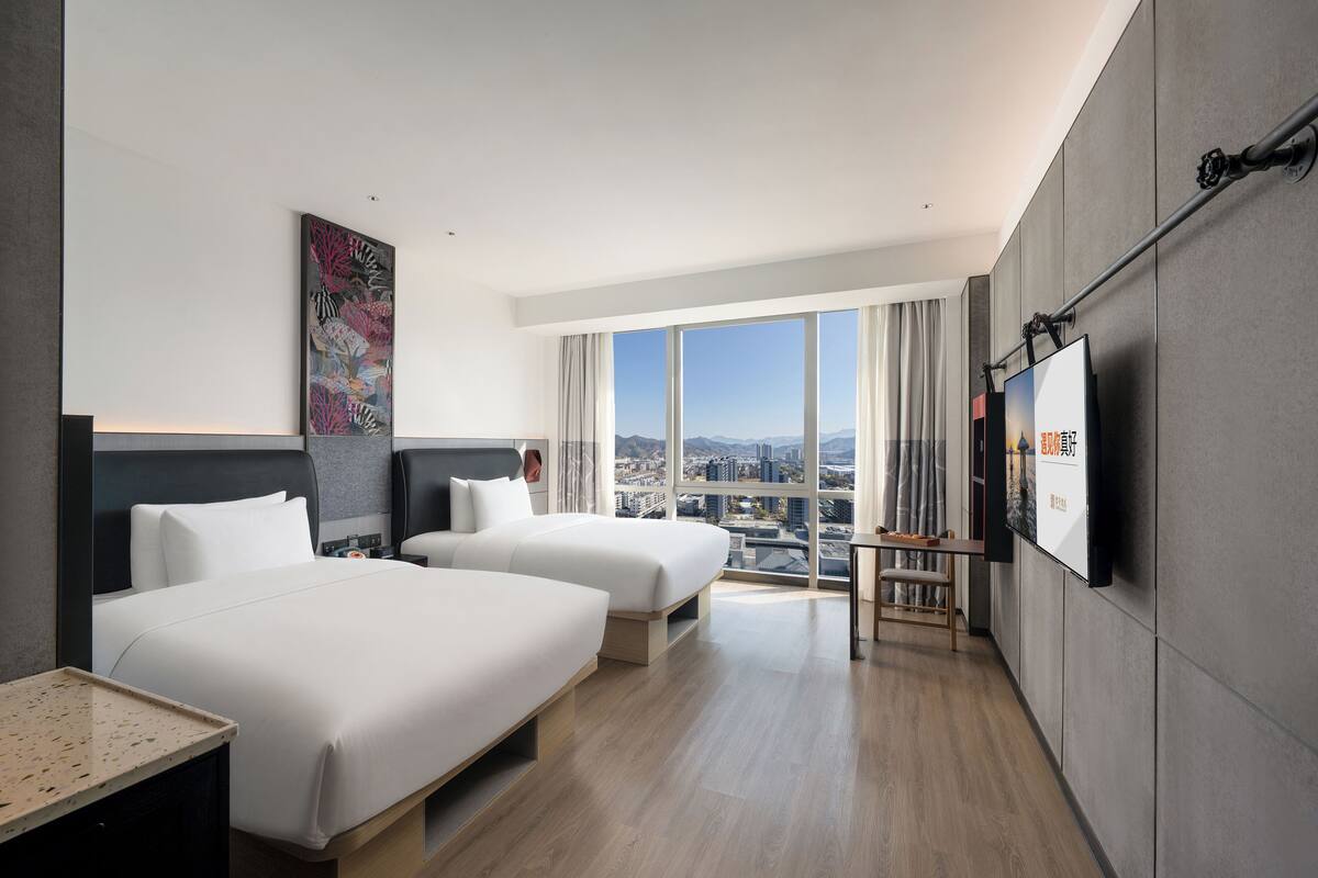 Habitación de hotel con dos camas, un ventanal con vistas a la ciudad, un televisor de pantalla plana y un cuadro en la pared.