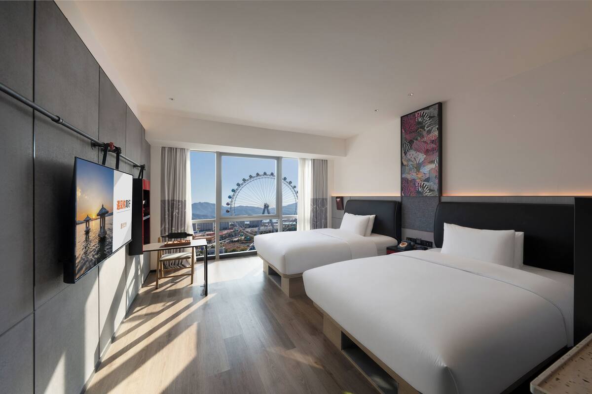 Habitación de hotel con dos camas, televisión, escritorio y ventana grande con vista a una noria.