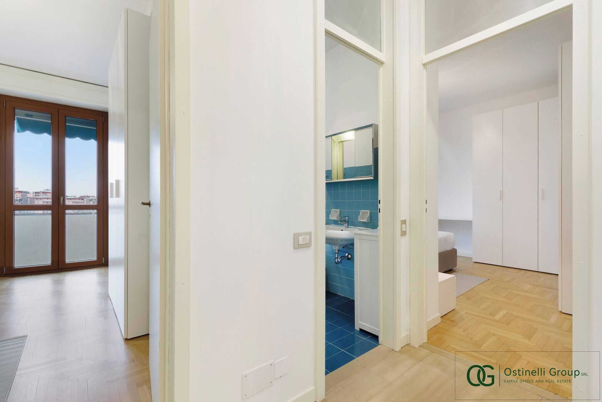 A modern apartment with a view through a glass door, a bathroom with blue tiles, and a wooden floor.