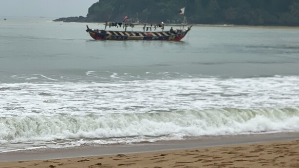 A boat with passengers on a calm sea, a sandy beach, and a forested hill in the background. - Simple and Eco friendly Beach Resort.Beach front bungalows , seafood ,vegetarian (Takoradi)