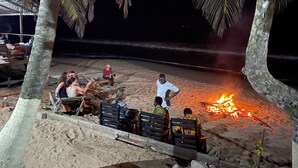 A beach scene with people sitting on benches and chairs around a campfire. - Simple and Eco friendly Beach Resort.Beach front bungalows , seafood ,vegetarian (Takoradi)