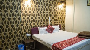 Un lit bien fait, recouvert d’une couvre-lit à motifs rouges et blancs, avec un oreiller et une couverture marron. Un seau bleu est posé sur le sol, à côté du lit, ainsi qu’une petite table de chevet en bois sur laquelle se trouve une lampe. Le mur est décoré d’un motif géométrique.