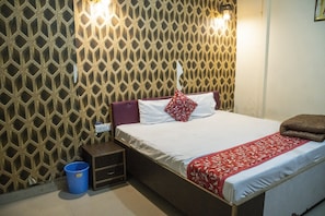 A neatly made bed with a red and white patterned bedspread, a pillow, and a brown blanket. There is a blue bucket on the floor beside the bed, and a small wooden nightstand with a lamp. The wall has a geometric patterned design. - PP GUEST HOUSE (Agra)