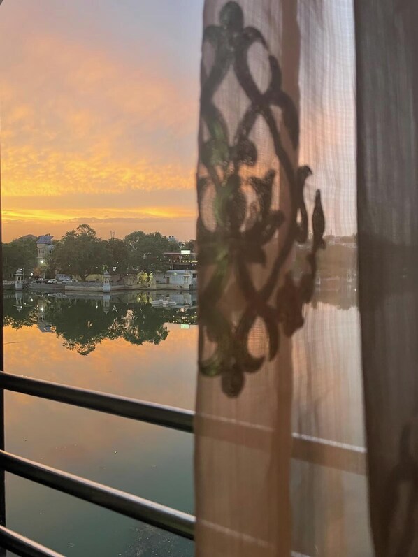 A view of a cityscape at sunset with buildings reflected in calm water, a decorative curtain partially obscuring the view, and a railing in the foreground. - Serene Lakeside Homestay (Udaipur)