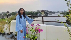 Eine Person steht auf einer Dachterrasse mit Blumentöpfen und blickt auf einen Fluss sowie Gebäude in der Ferne.