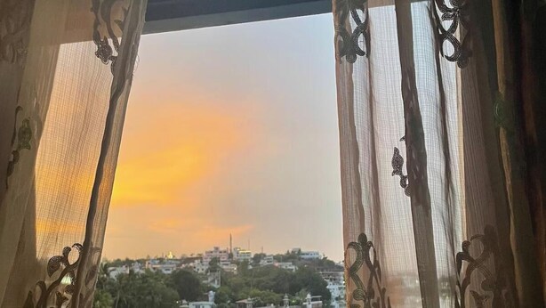 Blick durch ein Fenster mit aufwendigen Vorhängen, der ein ruhiges Gewässer und eine ferne Skyline im Abendrot reflektiert.
