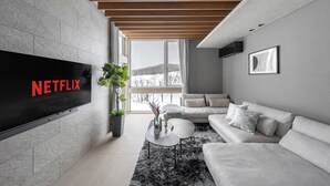 A modern living room with a flat-screen TV displaying "NETFLIX," a sofa, a coffee table, and a large window offering a view of the outdoors. - FURANO ECLAT (Furano)