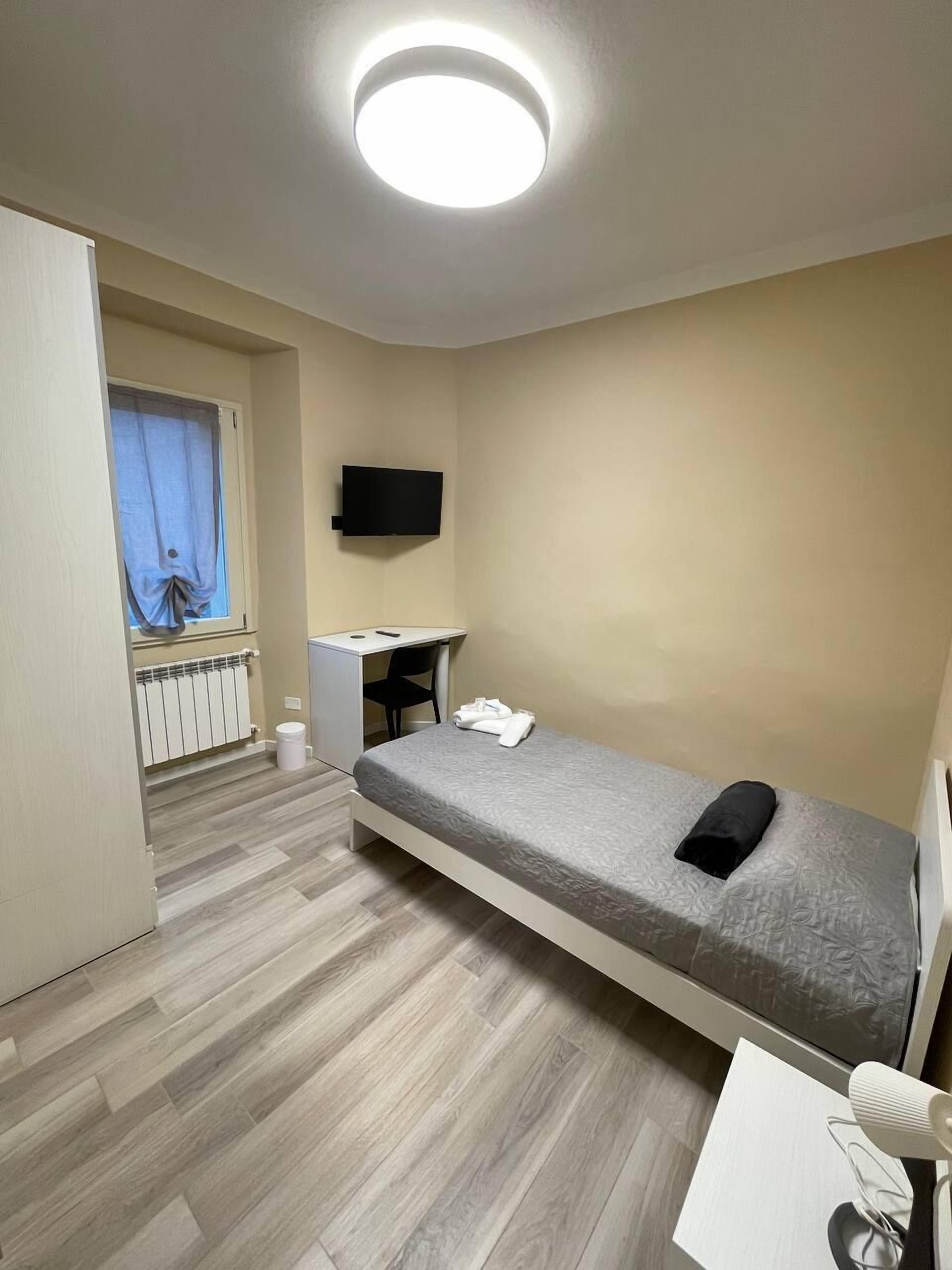 A small, modern hotel room with a single bed, a desk, a chair, a TV mounted on the wall, and a window with curtains.