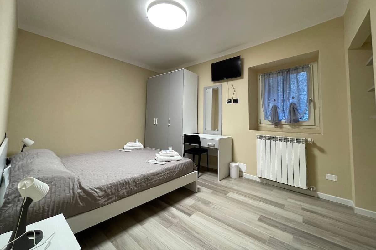 Une chambre à coucher avec un lit, un bureau, un miroir, une télévision et une fenêtre avec des rideaux.