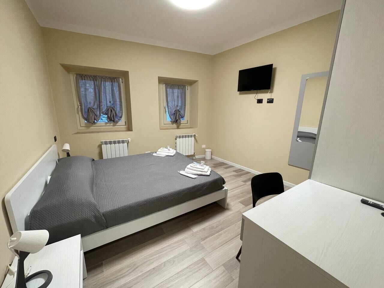 A hotel room with a bed, bedside tables, a desk, a chair, a TV, and two windows with curtains.