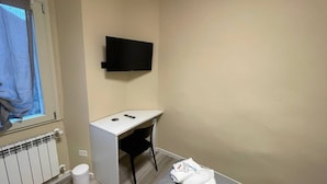Une petite pièce propre, équipée d’un bureau, d’une chaise, d’un téléviseur fixé au mur, d’un radiateur et d’une fenêtre avec un rideau.