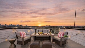Een terras op het dak met rieten meubels, een tafel gedekt voor twee personen, en een uitzicht op de skyline bij zonsondergang.