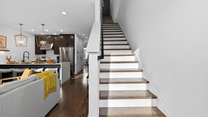 Een moderne keuken met een centraal eiland, een zithoek met een grijze bank en een geel plaid, en een trap met houten treden.