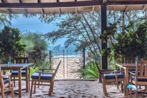 A beachside patio with wooden tables and chairs, a view of the ocean, and tropical plants. - Raman Resort (Calangute)