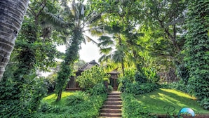 A garden pathway leading to a traditional building with a tiled roof, surrounded by lush greenery and palm trees.