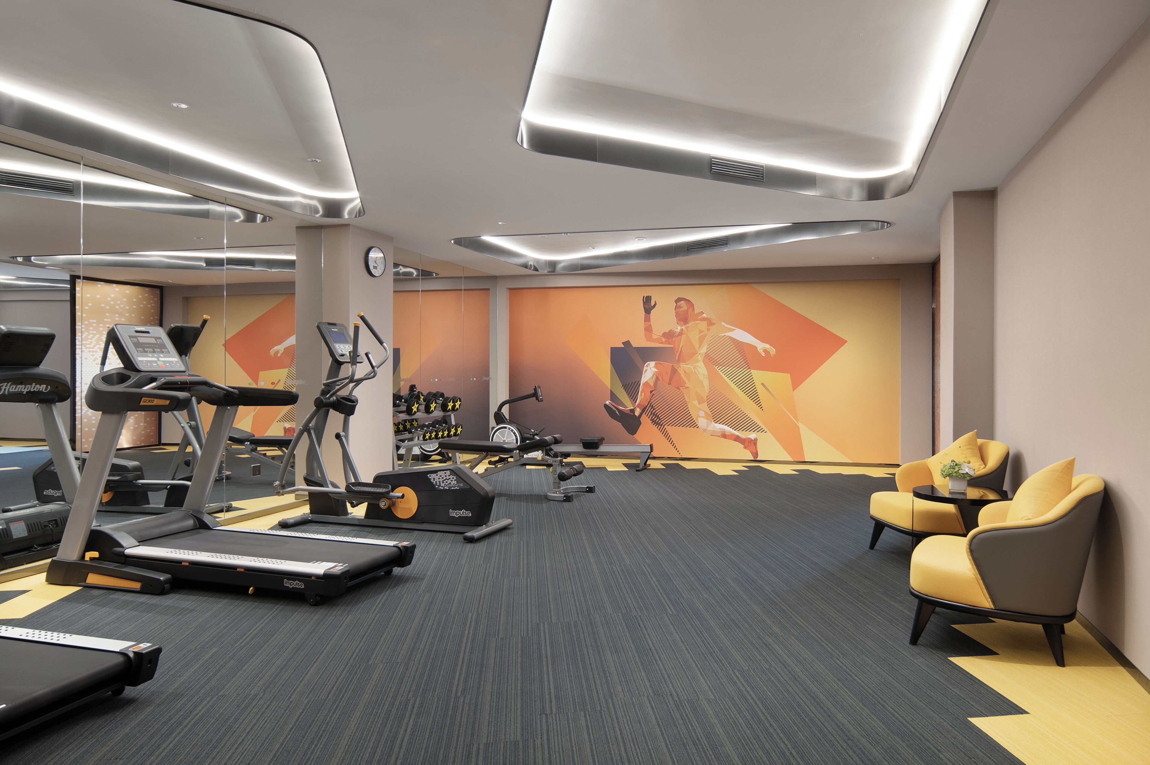 Ein modernes Fitnessstudio mit Laufbändern, Crosstrainern und Kraftstationen. Zwei gelbe Sessel und ein kleiner Tisch mit einer Pflanze sind ebenfalls vorhanden.