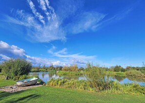 Un paysage paisible au bord d’un lac, avec un petit bateau amarré sur la rive, entouré d’une végétation luxuriante et sous un ciel bleu clair.
