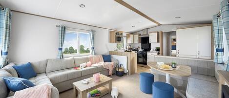 En moderne stue med sofa, sofabord og skamler, et køkkenområde med hårde hvidevarer, samt et spisebord.