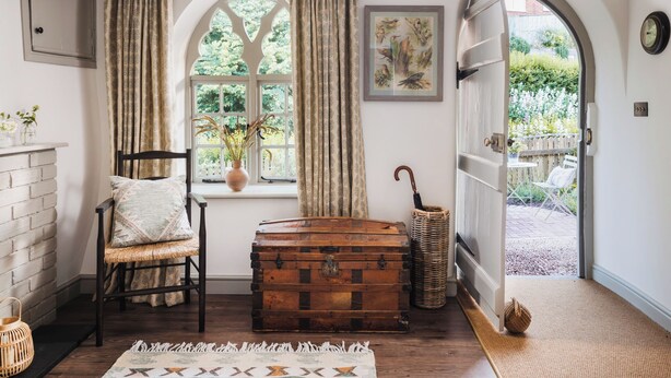 A room with a wooden chest, a wicker chair, a window with curtains, a framed picture, and an open door leading outside. - 3 bedroom accommodation in Budleigh Salterton (Budleigh Salterton)