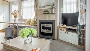 A modern living room with a fireplace, sofa, television, and a coffee table with a vase and a book. - This comfortable stylish caravan offers beautiful sea and country views. (Dorchester)