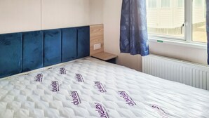 A bedroom with a bed, a window with curtains, a radiator, and a small wooden table. - This comfortable stylish caravan offers beautiful sea and country views. (Dorchester)