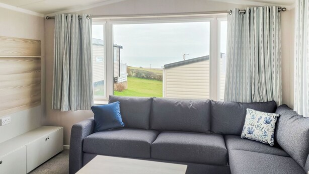 A modern living room with a grey sectional sofa, a small wooden coffee table, and a view of a house and greenery through large windows. - This comfortable stylish caravan offers beautiful sea and country views. (Dorchester)