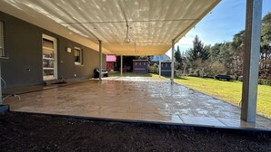 Ein überdachter Patio-Bereich mit Fliesenboden, Metallveranda und Blick auf einen Garten mit Rasen und Bäumen.