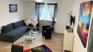 Ein modernes Wohnzimmer mit einem grauen Sofa, einem Glastisch und einem farbenfrohen Teppich. Ein schwarzer Mülleimer und ein weißes Schränkchen mit einem Fernseher sind ebenfalls vorhanden.