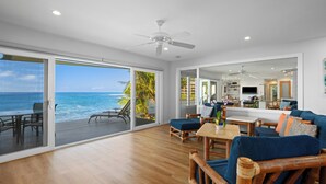 A spacious living area with a large sliding glass door leading to a balcony overlooking the ocean. There is a wooden deck, a seating area with blue cushions, and a kitchen visible through an open door. - Ho'ona Beach House by Alekona Kauai: 3BR Oceanfront Poipu Paradise (Koloa)