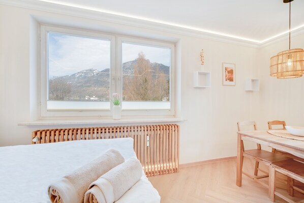 A bedroom with a large window, a bed with rolled towels, a wooden table, and a radiator.
