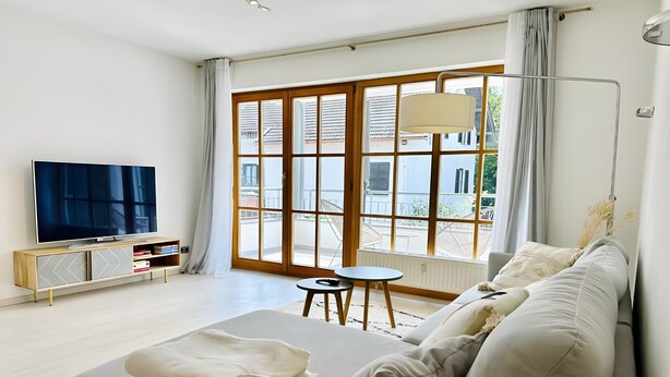 Een moderne woonkamer met een bank, een televisie, een klein tafeltje en grote ramen.