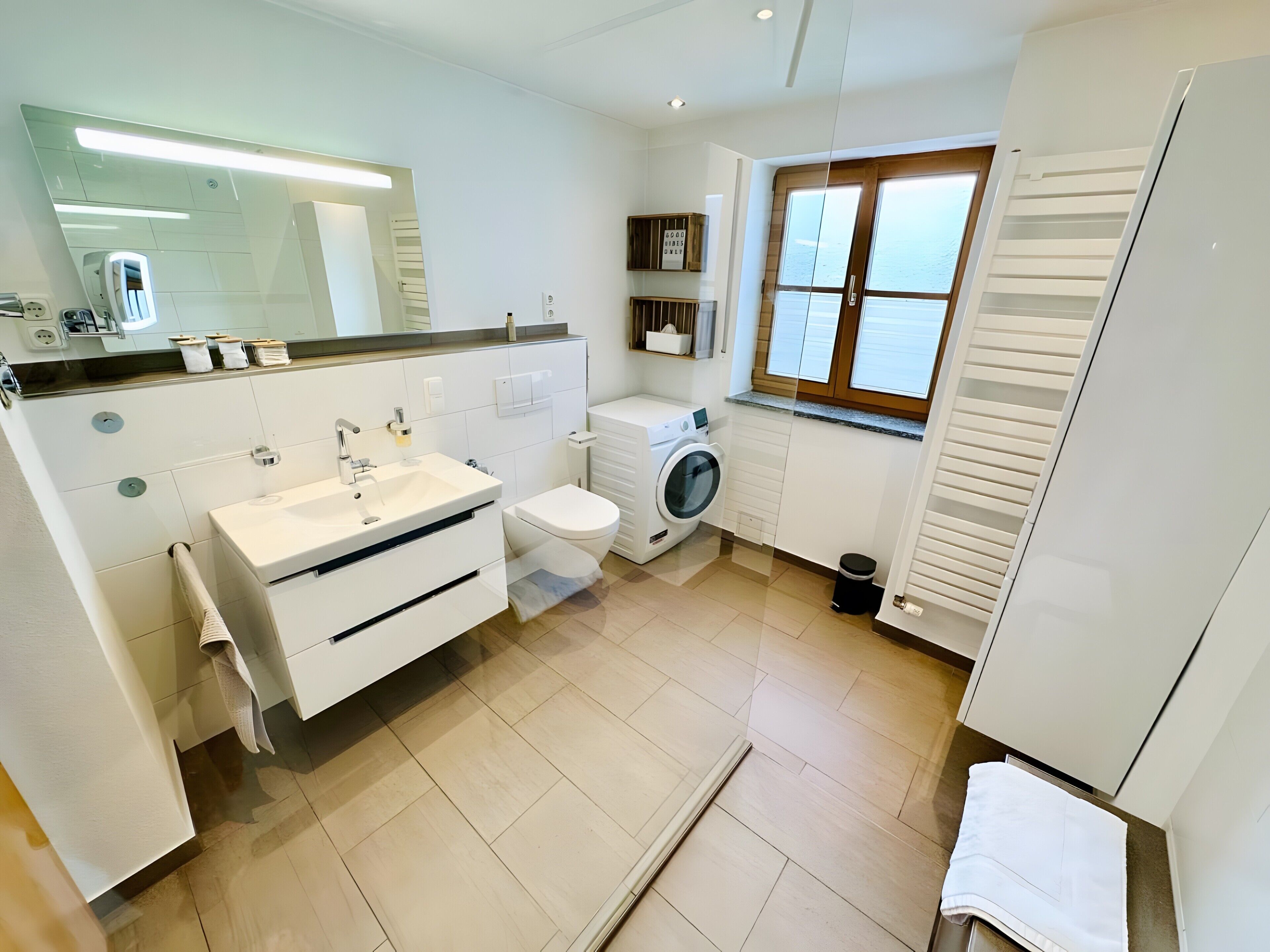 A modern bathroom with a sink, toilet, and washing machine, a mirror, and a window with blinds.