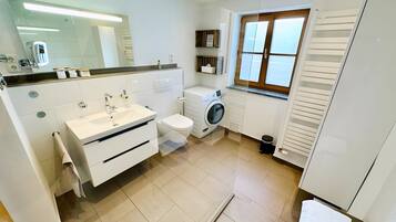 A modern bathroom with a sink, toilet, and washing machine, a mirror, and a window with blinds.