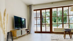 Een moderne woonkamer met een flatscreen televisie, een houten kast, een wit tapijt en een grote schuifdeur die uitkomt op een buitenruimte.