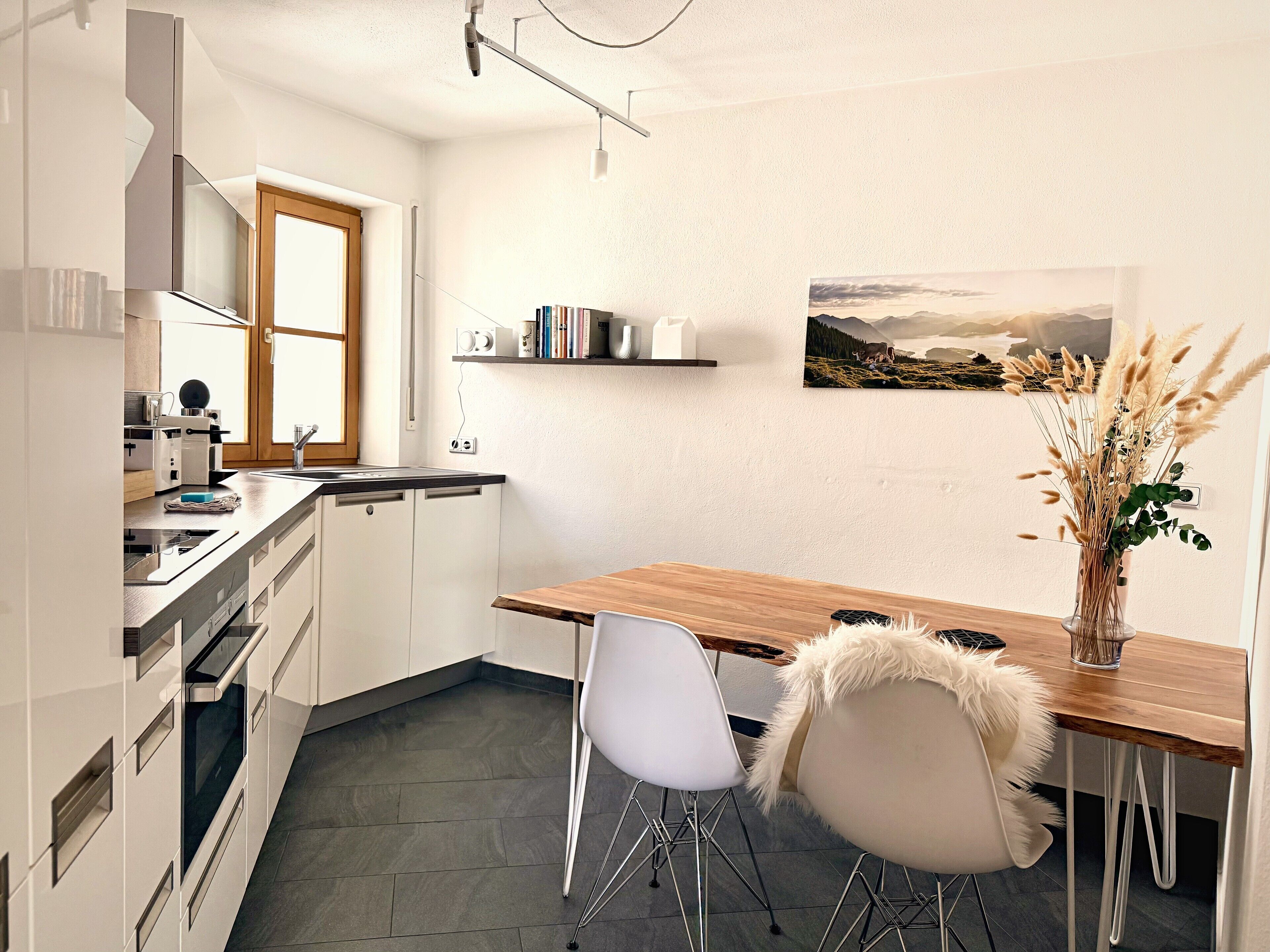 Una cocina moderna con armarios blancos, una mesa de comedor de madera y una silla blanca. Hay un cuadro en la pared y un jarrón con plantas secas sobre la mesa.
