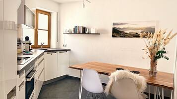 Una cocina moderna con armarios blancos, una mesa de comedor de madera y una silla blanca. Hay un cuadro en la pared y un jarrón con plantas secas sobre la mesa.