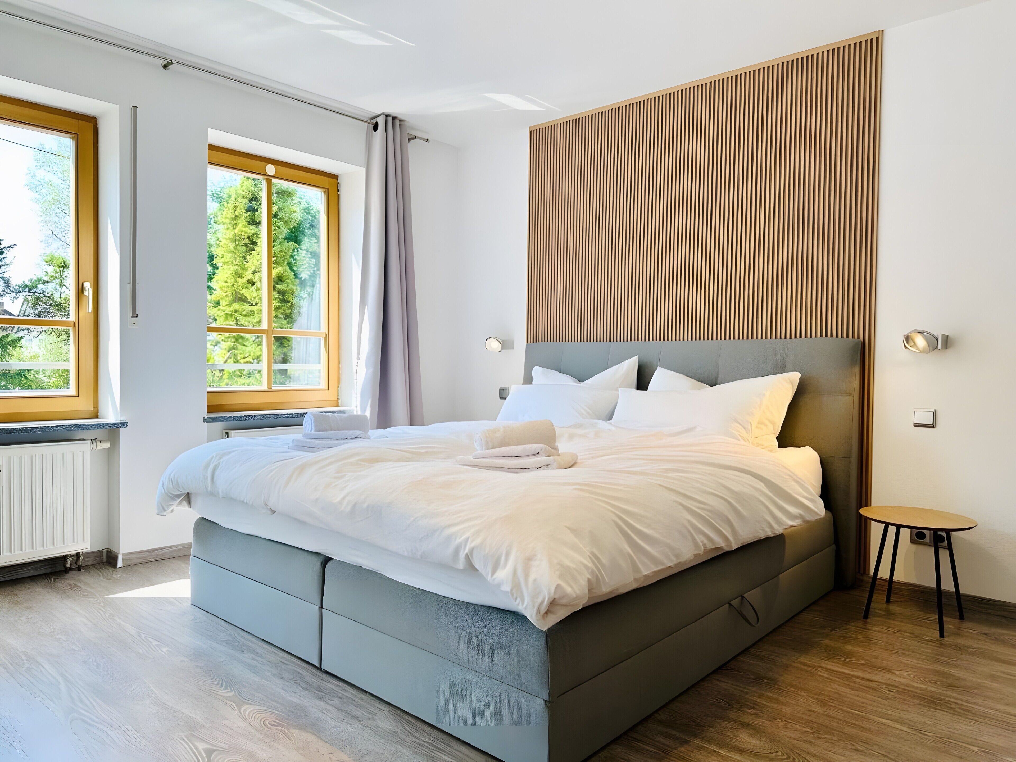 A modern bedroom with a large bed, a wooden panel wall, and a view of greenery outside.