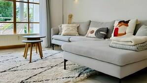Een moderne woonkamer met een grijze bank, een klein rond tafeltje en een groot raam met gordijnen.